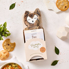 Close-up of a âSlumberkinsâ plush fox toy in its box, surrounded by cookies, rolls, greenery, and a bowl of soup partially visible nearby.