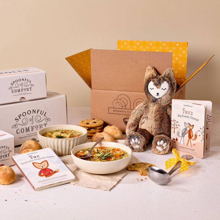 Comfort-themed gift set displayed with bowls of soup, rolls, cookies, Spoonful of Comfort boxes, a plush stuffed animal, childrenâs books, and a metal ladle with a yellow ribbon.