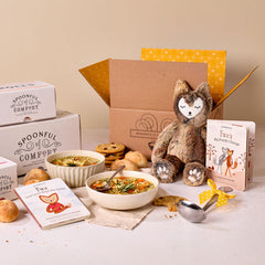 Comfort-themed gift set displayed with bowls of soup, rolls, cookies, Spoonful of Comfort boxes, a plush stuffed animal, childrenâs books, and a metal ladle with a yellow ribbon.