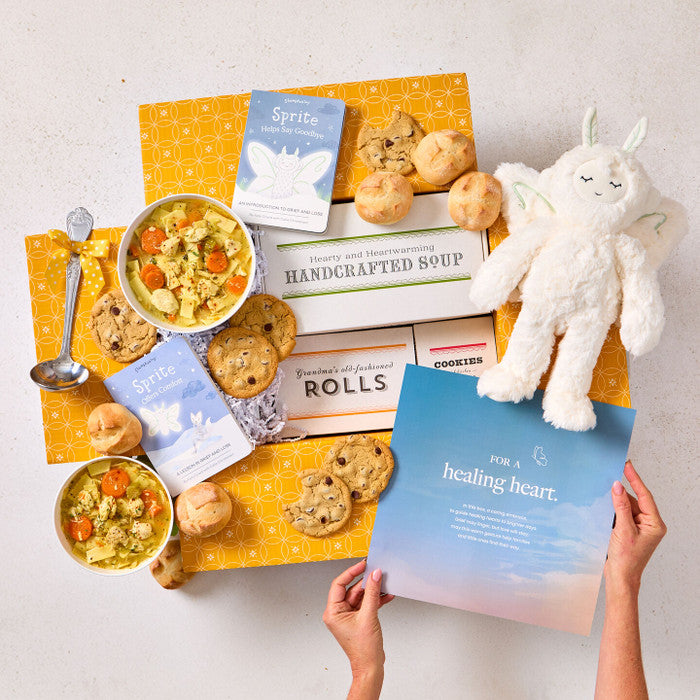 Overhead view of hands holding a blue card that reads âA Healing Heart,â next to a comforting gift arrangement with two bowls of soup, rolls, cookies, a box labeled âHandcrafted Soup,â a box of rolls, a plush stuffed animal, and a metal ladle with a yellow ribbon.