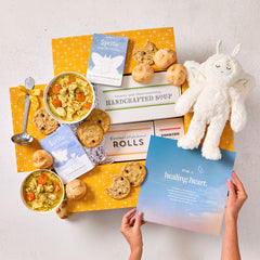 Overhead view of hands holding a blue card that reads âA Healing Heart,â next to a comforting gift arrangement with two bowls of soup, rolls, cookies, a box labeled âHandcrafted Soup,â a box of rolls, a plush stuffed animal, and a metal ladle with a yellow ribbon.