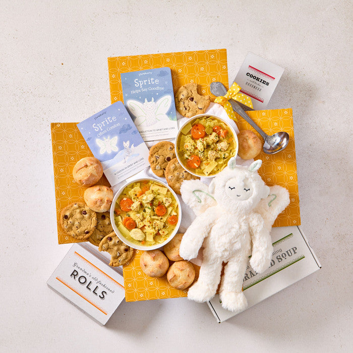 Overhead view of a comforting gift arrangement with two bowls of soup, rolls, cookies, a box labeled âHandcrafted Soup,â a box of rolls, a plush stuffed animal, childrenâs books, and a metal ladle with a yellow ribbon.