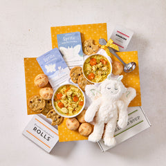 Overhead view of a comforting gift arrangement with two bowls of soup, rolls, cookies, a box labeled âHandcrafted Soup,â a box of rolls, a plush stuffed animal, childrenâs books, and a metal ladle with a yellow ribbon.