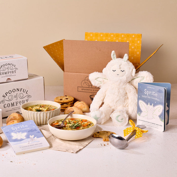 Comfort-themed gift set displayed with bowls of soup, rolls, cookies, Spoonful of Comfort boxes, a plush Sprite toy, childrenâs books, and a metal ladle with a yellow ribbon.