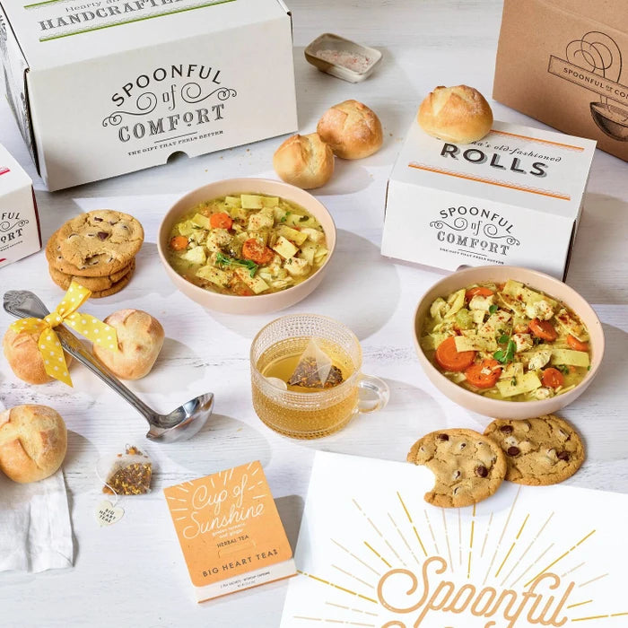 Soup gift set displayed with two bowls of soup, rolls, cookies, Spoonful of Comfort boxes, a glass of tea, a metal ladle with a yellow ribbon, and a card that reads âSpoonful of Sunshine.â