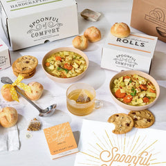 Soup gift set displayed with two bowls of soup, rolls, cookies, Spoonful of Comfort boxes, a glass of tea, a metal ladle with a yellow ribbon, and a card that reads âSpoonful of Sunshine.â