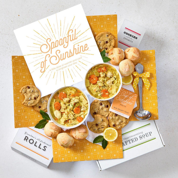 Overhead view of a yellow gift box arrangement with a bowl of soup, rolls, cookies, a container labeled âHandcrafted Soup,â a metal ladle with a yellow ribbon, lemon slices, a package of tea sachets, and a card that reads âSpoonful of Sunshine.â
