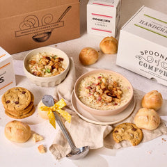 Soup gift set displayed with two bowls of soup topped with garnish, rolls, cookies, Spoonful of Comfort boxes, and a metal ladle with a yellow ribbon arranged on a table.