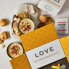 Overhead view of an open yellow gift box with a card that reads âLove by the Spoonful,â surrounded by bowls of soup, rolls, cookies, and packaged items.