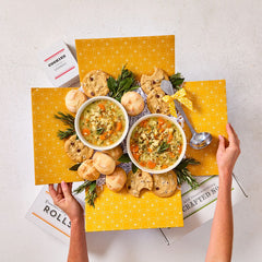 Overhead view of hands holding an open yellow gift box filled with two bowls of soup, rolls, cookies, greenery, and a metal ladle.