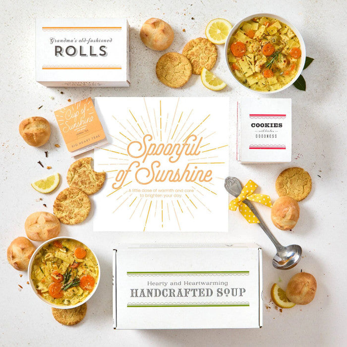 Overhead view of a sunshine-themed gift arrangement with two bowls of soup, rolls, cookies, lemon slices, a box labeled âHandcrafted Soup,â a box of rolls, a box of cookies, a metal ladle with a yellow ribbon, and a card that reads âSpoonful of Sunshine.â