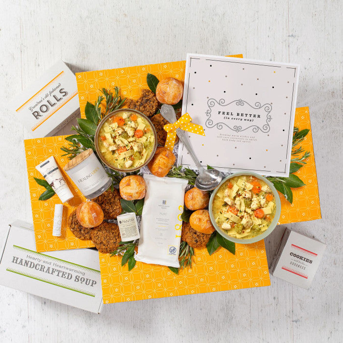 Overhead view of a yellow gift box arrangement with two bowls of soup, rolls, cookies, a container labeled âHandcrafted Soup,â a metal ladle with a yellow ribbon, a candle, small self-care items, and a card.