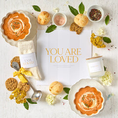 Overhead view of a comforting gift arrangement with two bowls of tomato soup, rolls, cookies, a candle, cozy socks labeled âI Am Loved,â a metal ladle with a yellow ribbon, greenery, and an open card that reads âYou Are Loved.â