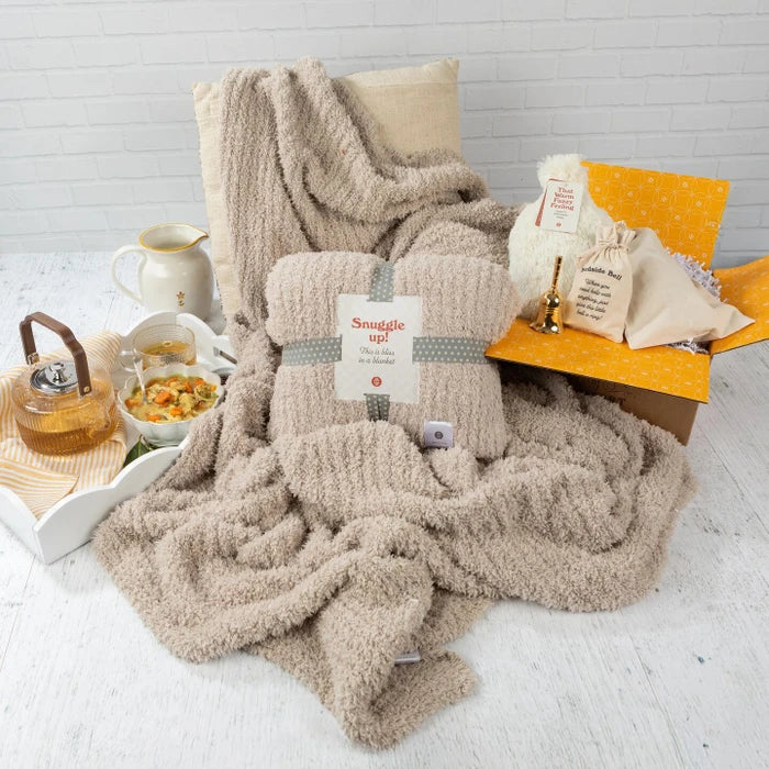 Cozy get-well-themed arrangement featuring a plush throw blanket with a âSnuggle Upâ pack on top, alongside a tray with soup, tea, and honey, plus a âBedside Bellâ and self-care items displayed on a yellow box.