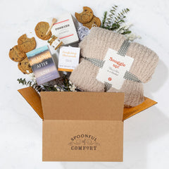 Open Spoonful of Comfort gift box filled with a cozy knit blanket labeled âSnuggle Up,â cookies, a box of cookies, a package of chamomile tea, a jar of honey with a dipper, and a book titled âHealing After Loss,â arranged with greenery.