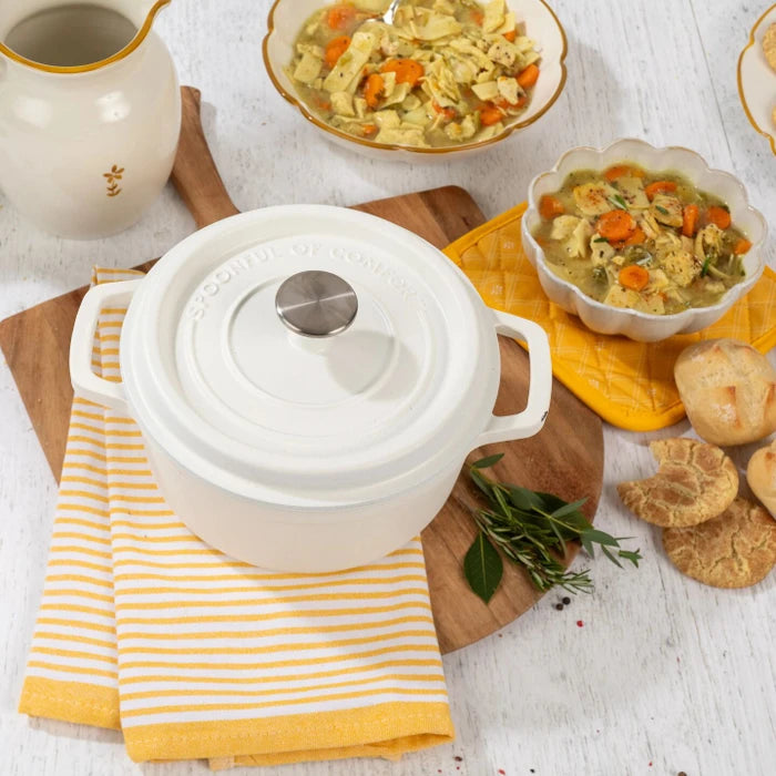 Overhead view of a white Dutch oven with lid on a yellow striped towel, with bowls of soup, rolls, cookies, and herbs arranged around it.