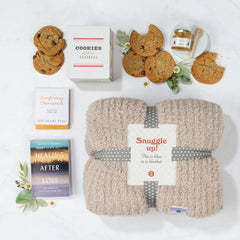 Overhead view of a comfort-themed gift arrangement with a cozy knit blanket labeled âSnuggle Up,â cookies, a box of cookies, a candle, a jar of honey with a dipper, and a book titled âHealing After Loss,â arranged with greenery.