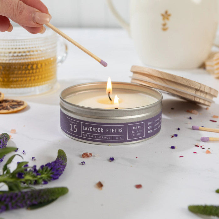Close-up of a hand lighting a âLavender Fieldsâ candle with a match, surrounded by scattered lavender, matches, and a glass of tea.