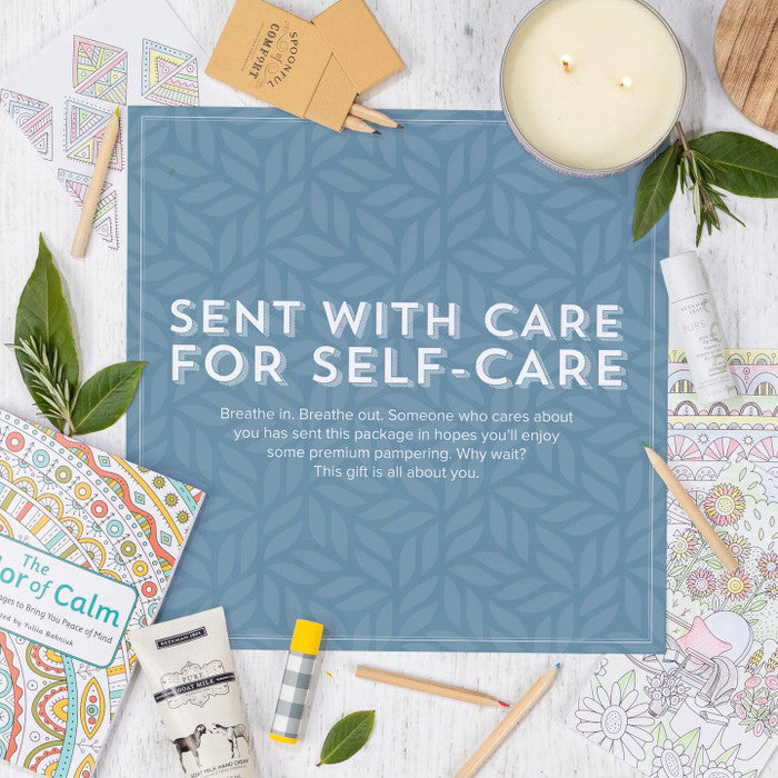 Flat lay of a self-care arrangement with a candle, coloring book, pencils, tea, and small items surrounding a blue card that reads âSent With Care for Self-Care.â