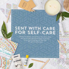 Flat lay of a self-care arrangement with a candle, coloring book, pencils, tea, and small items surrounding a blue card that reads âSent With Care for Self-Care.â