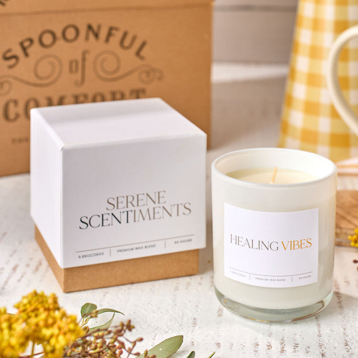 Close-up of a âHealing Vibesâ candle next to its box labeled âSerene Scents,â with dried flowers and a Spoonful of Comfort box in the background.