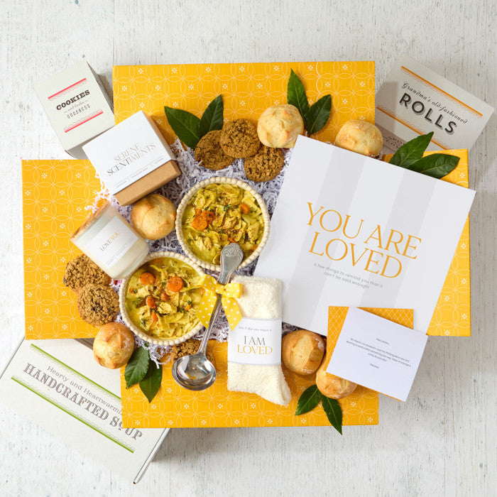 Overhead view of a yellow gift box arrangement with two bowls of soup, rolls, cookies, a container labeled âHandcrafted Soup,â a metal ladle, a candle, a pair of cozy socks, and a card that reads âYou Are Loved.â