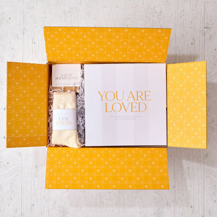 Open yellow gift box displaying a card that reads âYou Are Lovedâ alongside a pair of cozy socks labeled âI Am Loved.â