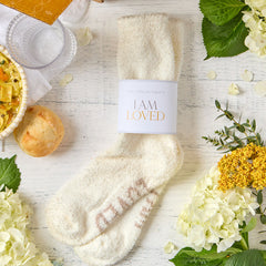 Close-up of a pair of cozy cream-colored socks labeled âI Am Loved,â surrounded by flowers, greenery, and nearby food items.