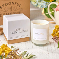 Close-up of a âLove Youâ candle being lit next to its box labeled âSerene Scents,â with dried flowers and a Spoonful of Comfort box in the background.