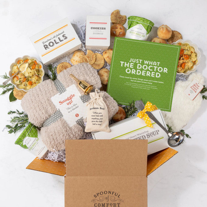 Open Spoonful of Comfort gift box filled with a get-well-themed arrangement including two bowls of soup, rolls, cookies, containers of soup with green lids, a cozy throw blanket, a âWarm Fuzziesâ warm/cool pack, a âBedside Bellâ for assistance, a metal ladle with a yellow ribbon, greenery, and a green card that reads âJust What the Doctor Ordered.â