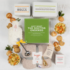 Overhead view of a get-well-themed gift arrangement with two bowls of soup, rolls, cookies, containers labeled âHandcrafted Soup,â a cozy throw blanket, a âWarm Fuzziesâ warm/cool pack, a âBedside Bell,â a metal ladle with a yellow ribbon, greenery, and a green card that reads âJust What the Doctor Ordered.â
