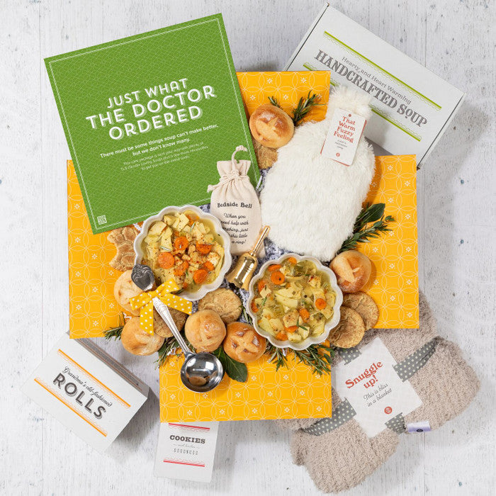 Overhead view of a yellow gift box filled with two bowls of soup, rolls, cookies, a container of handcrafted soup, a cozy throw blanket, a Warm Fuzzies warm/cool pack, and a green card that reads âJust What the Doctor Ordered.â