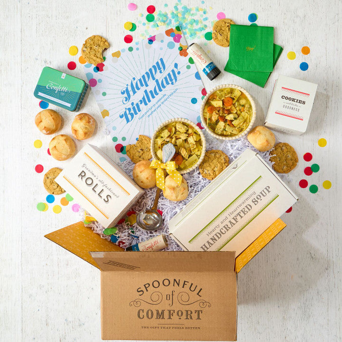Open Spoonful of Comfort gift box with a birthday-themed arrangement including two bowls of soup, rolls, cookies, colorful confetti, green napkins, confetti poppers, packaged items, and a card that reads âHappy Birthday!â
