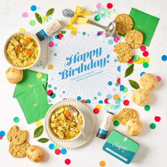 Overhead view of a birthday-themed gift arrangement with two bowls of soup, rolls, cookies, a container labeled âHandcrafted Soup,â a metal ladle with a yellow ribbon, green birthday napkins, confetti poppers, colorful confetti, and a card that reads âHappy Birthday!â