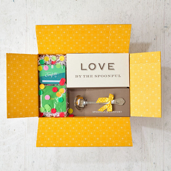 Open yellow gift box displaying a card that reads âLove by the Spoonful,â a metal ladle with a yellow ribbon, and two confetti poppers with colorful confetti packaging.