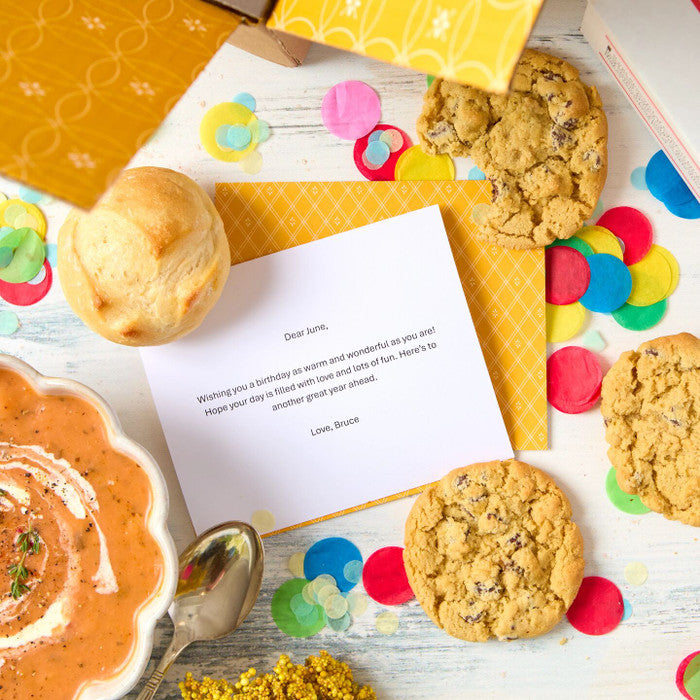 Close-up of a birthday card placed among a bowl of soup, rolls, cookies, and colorful confetti on a yellow background.