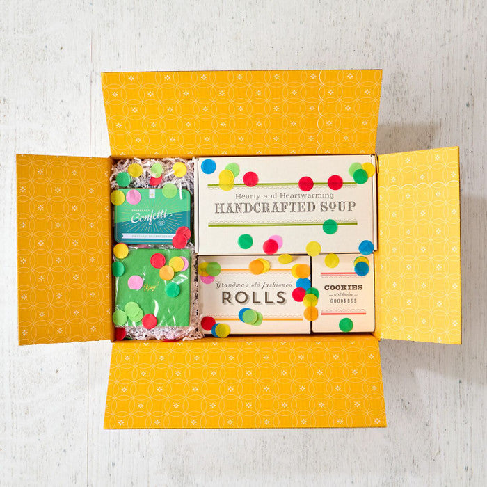 Open yellow gift box filled with packages labeled âHandcrafted Soup,â âRolls,â and âCookies,â alongside two confetti poppers and scattered colorful confetti.