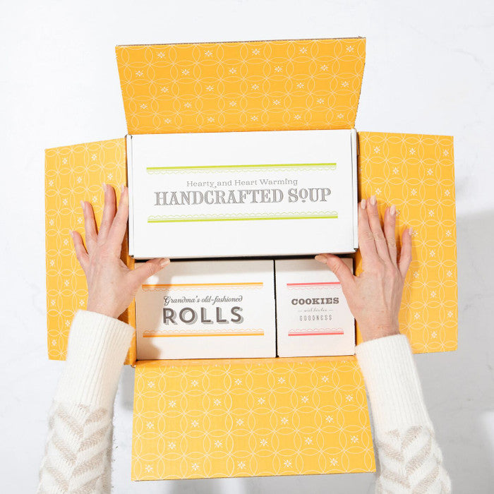 Overhead view of hands opening a yellow gift box containing packages labeled âHandcrafted Soup,â âRolls,â and âCookies.â