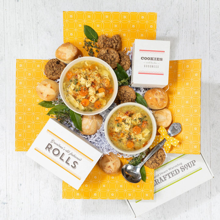 Overhead view of a yellow gift box arrangement with two bowls of chicken soup, rolls, cookies, a box of cookies, a box of rolls, a container labeled âHandcrafted Soup,â and a metal ladle arranged with greenery.