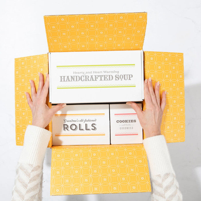 Top view of hands opening a yellow gift box containing packaged rolls, a container labeled handcrafted soup, and a box of cookies.