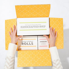 Top view of hands opening a yellow gift box containing packaged rolls, a container labeled handcrafted soup, and a box of cookies.