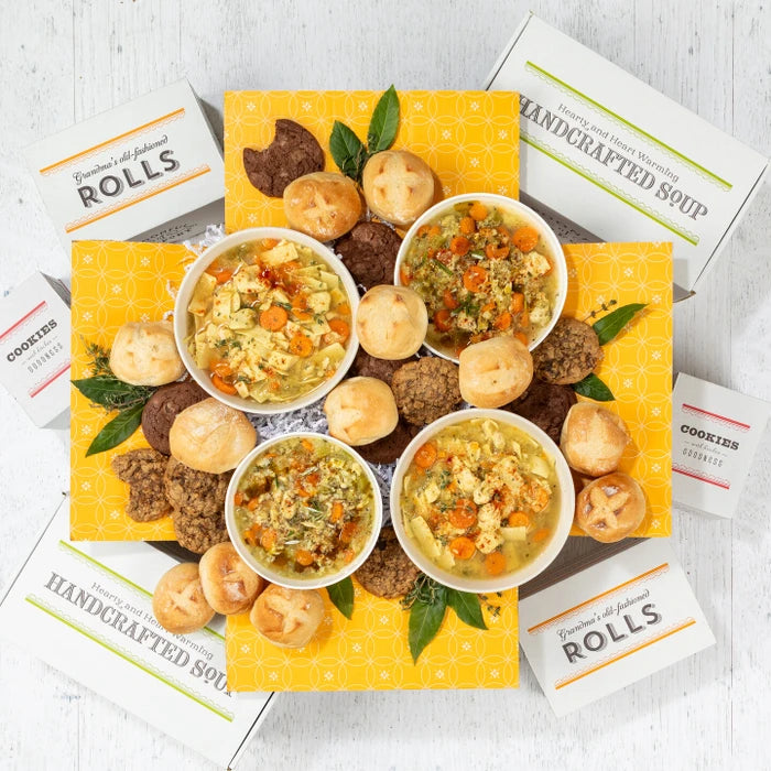 Overhead view of a large soup gift arrangement with four bowls of soup, rolls, cookies, multiple boxes labeled âHandcrafted Soupâ and âRolls,â and greenery arranged on a yellow box.