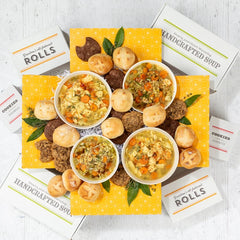Overhead view of a large soup gift arrangement with four bowls of soup, rolls, cookies, multiple boxes labeled âHandcrafted Soupâ and âRolls,â and greenery arranged on a yellow box.
