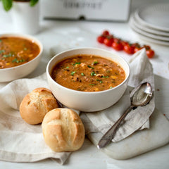 Two bowls of soup on a table with bread rolls and a spoon beside them.