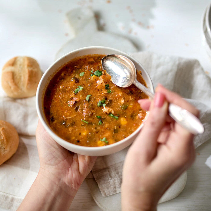 Hands holding a bowl of soup with herbs, with bread rolls nearby.