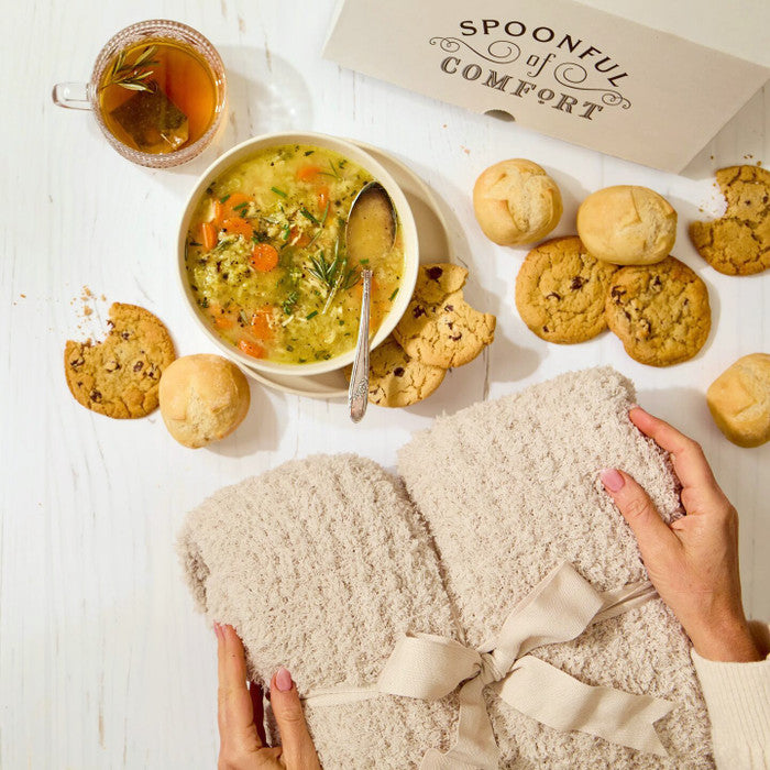 Overhead view of hands holding a cozy knit blanket next to a bowl of soup, rolls, cookies, and a Spoonful of Comfort box on a light surface.