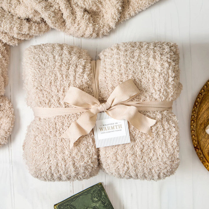 Close-up of a soft, plush knit blanket tied with a ribbon and labeled âWarmth,â folded on a light surface.