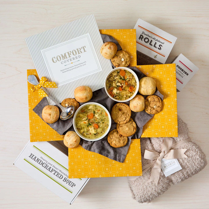 Overhead view of a comfort-themed gift arrangement with two bowls of soup, rolls, cookies, a box labeled âHandcrafted Soup,â a box of rolls, a metal ladle with a yellow ribbon, a cozy knit blanket, and a card that reads âComfort Covered.â
