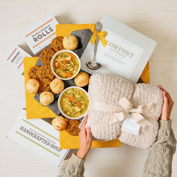 Overhead view of hands holding a cozy knit blanket next to a comfort-themed gift arrangement with two bowls of soup, rolls, cookies, a box labeled âHandcrafted Soup,â a metal ladle with a yellow ribbon, and a card that reads âComfort Covered.â