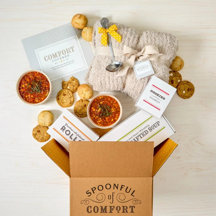 Open Spoonful of Comfort gift box filled with a comfort-themed arrangement including two bowls of soup, rolls, cookies, a box labeled âHandcrafted Soup,â a box of rolls, a metal ladle with a yellow ribbon, a cozy knit blanket, and a card that reads âComfort Covered.â